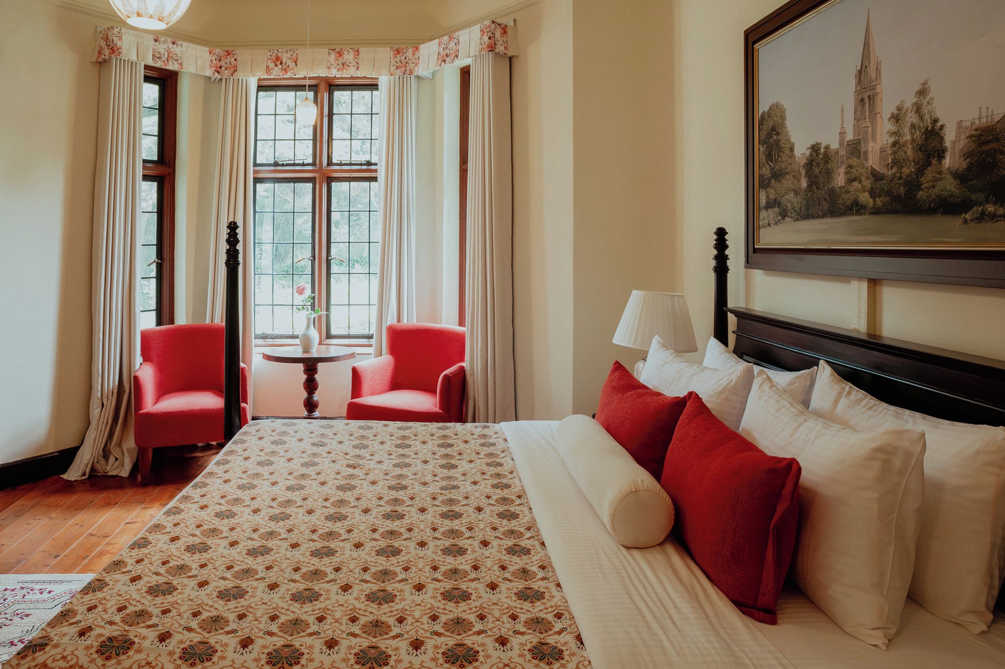 A hotel room with a large bed, two red chairs, a small table, and a painting on the wall.