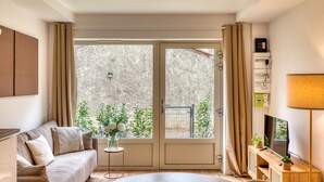 Una sala moderna con un sofá, una mesita auxiliar con un florero, un televisor sobre un mueble de madera y un ventanal con cortinas.