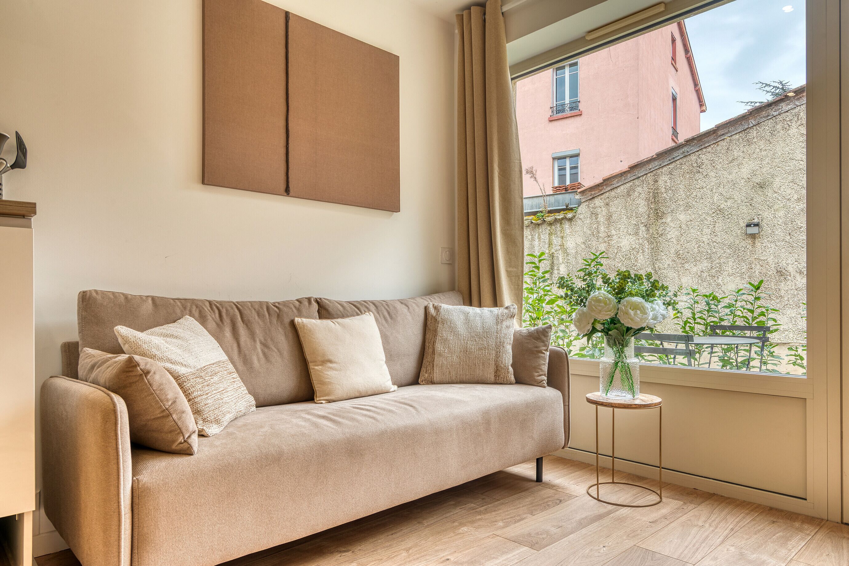 A beige sofa with cushions, a small round table with a vase of flowers, and a window with a view of a building and greenery.