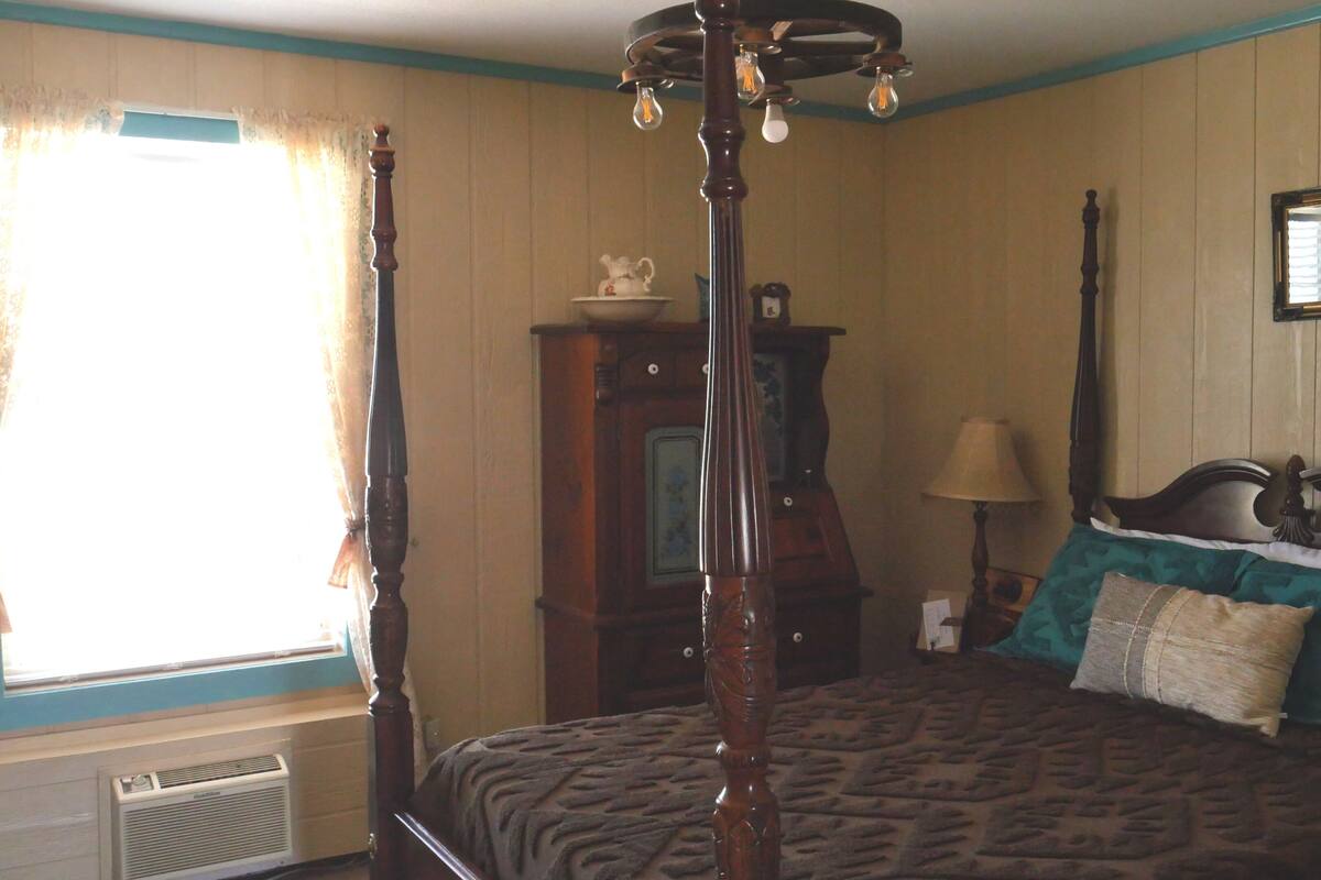 A room with a four-poster bed, a wooden dresser, a mirror on the wall, and a window with curtains.