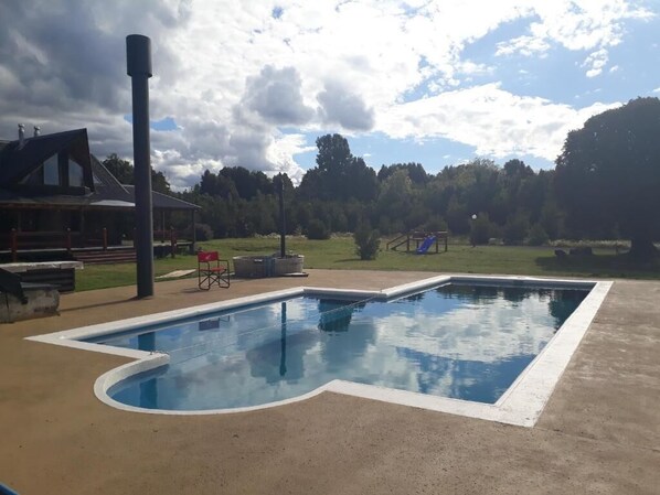 A swimming pool with a concrete deck, a wooden deck chair, and a small red chair. - Martina Bungalows Country Lodge (Puerto Varas)