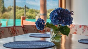 Una mesa de madera con un jarrón de flores azules, vista de montañas a través de amplios ventanales y un techo de madera.