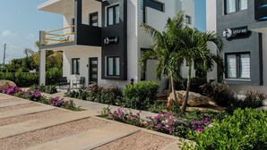 A modern two-story building with a balcony, a sign reading "MEADS BAY," and a well-maintained garden with various plants and flowers. - Genesis Residences 
Meads Bay 1 - Downstairs Unit  (The Cove)
