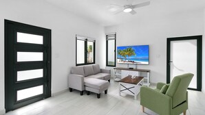 A modern living room with a grey sofa, a green armchair, a coffee table, and a TV displaying a beach scene. - Genesis Residences 
Meads Bay 1 - Downstairs Unit  (The Cove)
