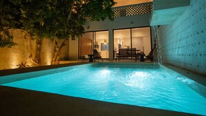Una piscina con caída de agua, rodeada por un edificio con puertas de vidrio y un área de patio.