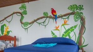 A bed with a blue bedspread and a wooden headboard, set against a wall with a mural of a tree branch and tropical plants. - Ecolodge Kala Luna Resort (Drake Bay)