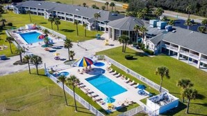 Vista aérea de un resort con múltiples piscinas, tumbonas y un edificio grande con techo curvo.