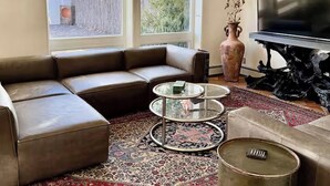 Een woonkamer met een bruine leren hoekbank, een glazen salontafel, een ronde metalen tafel, een vloerkleed met dessin, een vaas met gedroogde bloemen en een flatscreen-televisie.