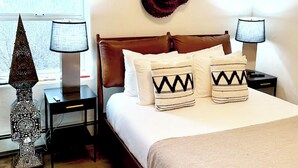 Een net opgemaakt bed met gestreepte kussens, twee bedlampjes, een decoratieve wandhanger en een bijzondere vloerlamp.