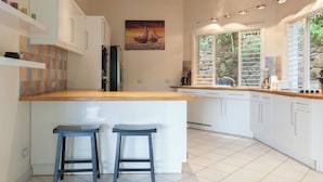 A modern kitchen with white cabinets, a wooden countertop, two bar stools, a painting of a sunset, and a stone wall behind the sink. - Villa Tramonto. Great view, pool, exclusive area and beach at walking distance. (St. Johns)