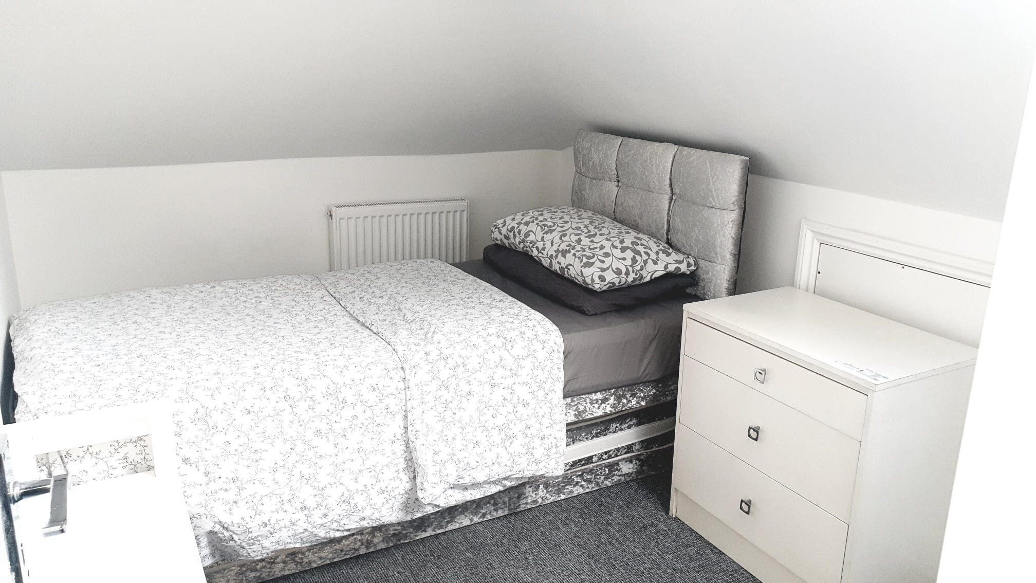A bedroom with a bed, a bedside table, a radiator, and a dresser.