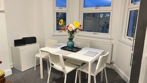 Une pièce avec une table blanche, quatre chaises blanches, un vase de fleurs et deux fenêtres donnant sur la ville.