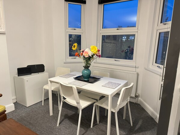 Una habitación con una mesa blanca, cuatro sillas blancas, un jarrón con flores y dos ventanas con vistas a la ciudad.