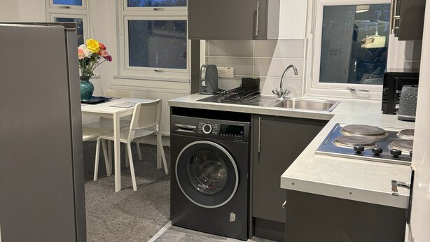 Une cuisine compacte dotée d’un lave-linge intégré, un coin repas avec une table et des chaises blanches, et de grandes fenêtres offrant une vue sur l’extérieur.