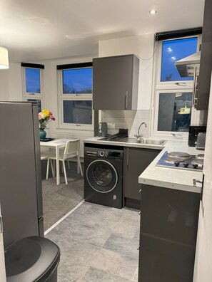 Una cocina compacta con lavadora integrada, una zona de comedor con mesa y sillas blancas, y ventanas amplias que ofrecen vistas al exterior.