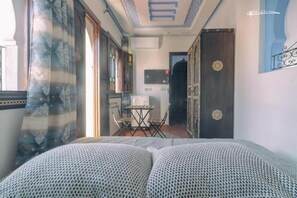 A room with a bed, a small table, and a chair. There is a window with curtains, a door with a decorative handle, and a wall with a framed picture. - Dar Rafik (Chefchaouen)