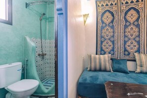 A bathroom with a glass shower, a toilet, and a bed with patterned pillows. - Dar Rafik (Chefchaouen)