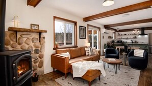 A cozy living room with a stone fireplace, a leather sofa, and a dining area with a kitchen in the background. - Aspen Glow Cabin - a charming 2-bedroom cabin with mountain views & wood stove (Bailey)