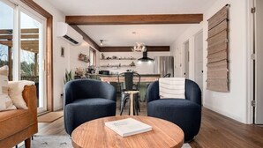 A modern living room with a round wooden coffee table, two blue armchairs, and a sofa. A kitchen is visible in the background with a dining table and chairs. - Aspen Glow Cabin - a charming 2-bedroom cabin with mountain views & wood stove (Bailey)