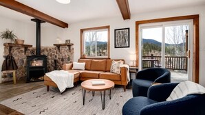 A cozy living room with a stone fireplace, a sofa, a coffee table, and a view of the outdoors through large windows. - Aspen Glow Cabin - a charming 2-bedroom cabin with mountain views & wood stove (Bailey)