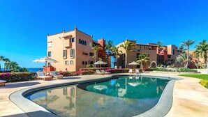 Un espace piscine doté de chaises longues et de parasols, entouré de palmiers et bordé d’un immeuble avec de nombreux balcons.