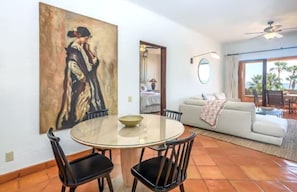 A dining area with a round table and chairs, a painting on the wall, a sofa, and a view of the outdoors through a large window. - 2BD Beach Front Luxury in Cabo (San José del Cabo)