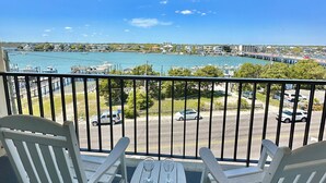 Ein Balkon mit zwei weißen Schaukelstühlen und einem kleinen Tisch, mit Blick auf eine Marina mit Booten und blauem Himmel.