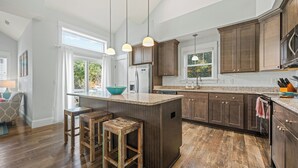 A kitchen with wooden cabinets, a large island with stools, pendant lighting, and a view of the outdoors. - 4BR All Ensuite Baths + Bonus Living Area + Hot Tub + Pet-Friendly + Corolla Lt (Corolla)