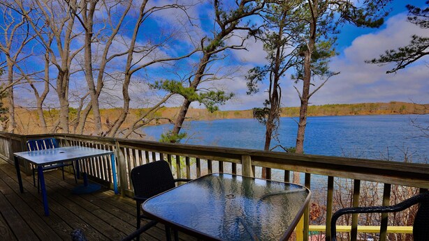 A wooden deck with a table and chairs, overlooking a body of water and trees.