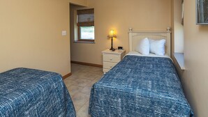 A bedroom with two beds, a nightstand with a lamp, a window with blinds, and a small picture on the wall.