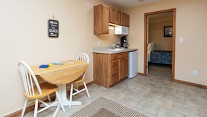Une cuisine compacte avec des meubles en bois, un plan de travail blanc et un micro-ondes encastré. Une table à manger avec des chaises est placée devant la cuisine.