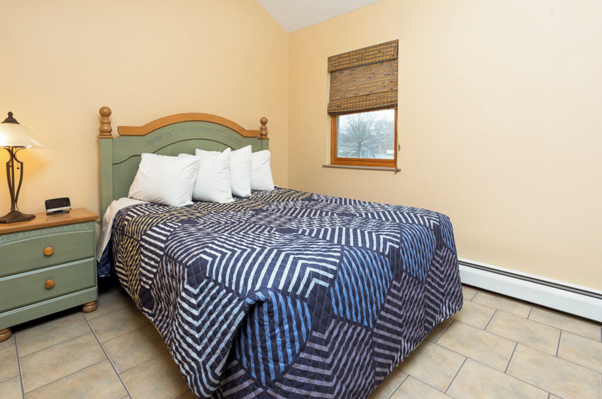A bedroom with a bed, a nightstand, a lamp, and a window with blinds.