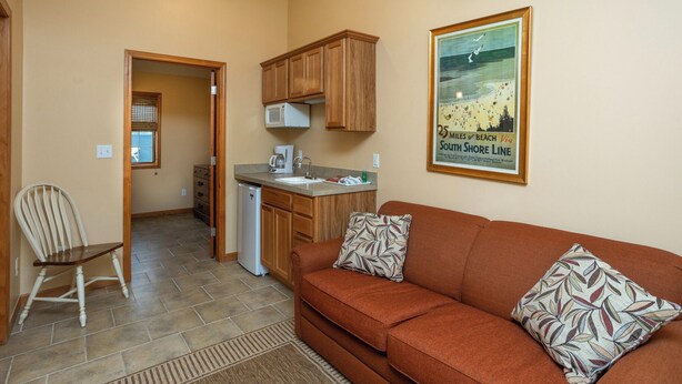 A compact living space with a kitchenette, a brown sofa with patterned cushions, and a vintage beach poster on the wall. - A Suite Place .5 mi from Beach (Michigan City)