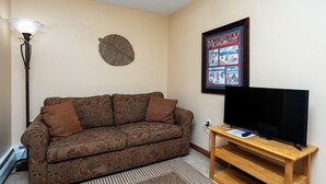 Un salon avec un canapé à motifs, un téléviseur à écran plat posé sur un meuble en bois, une lampe sur pied et un tableau encadré accroché au mur.