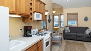 A compact kitchen with wooden cabinets, a microwave, a gas stove, and a coffee maker. There's a dining area with a table and chairs, and a gray sofa in the living room. - A Suite Place .5 mi from Beach (Michigan City)