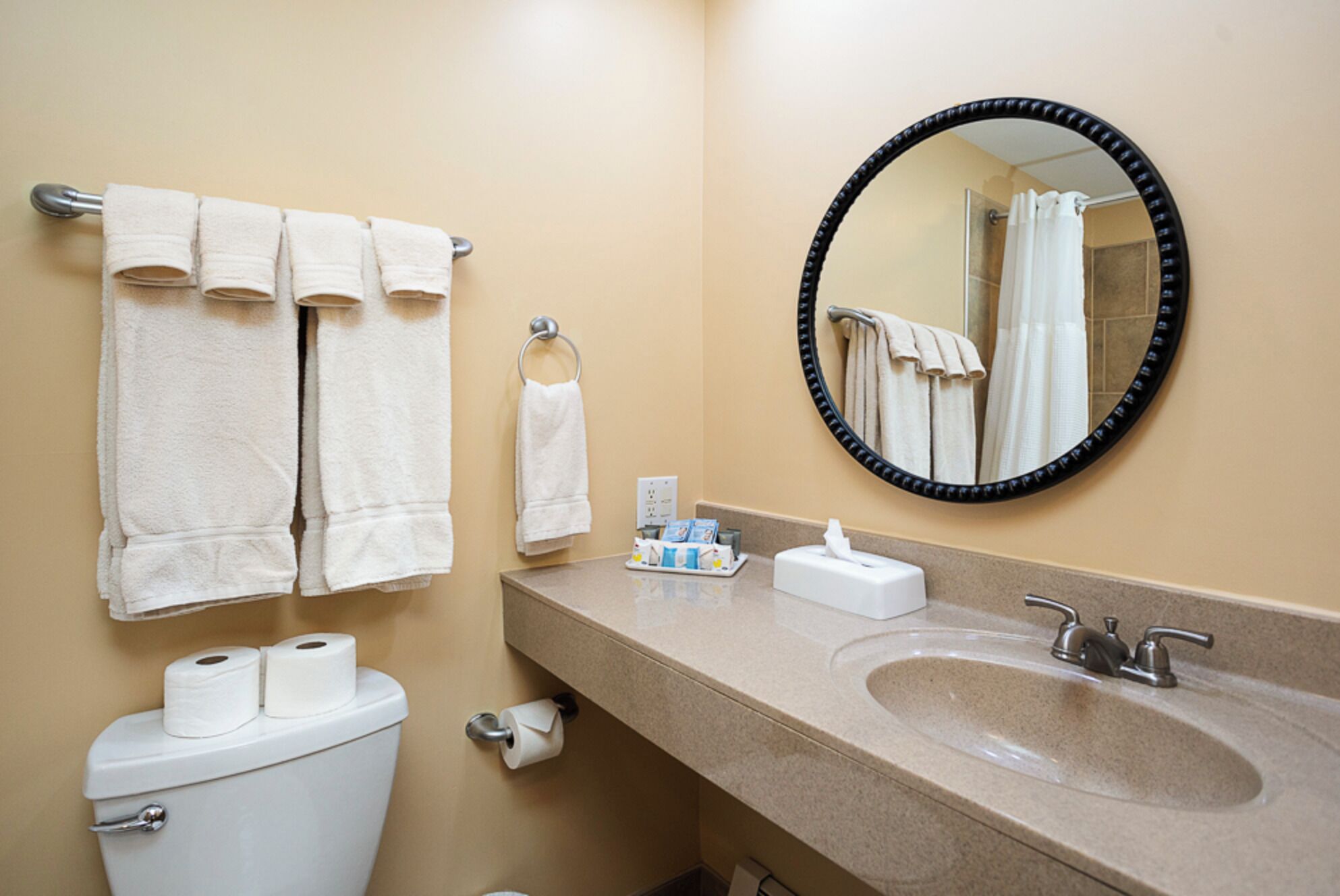 A bathroom with a toilet, sink, and mirror. There are towels hanging on a rack and a towel dispenser.