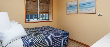 A small, neatly arranged bedroom with a bed, a window with blinds, a ceiling fan, and two wall-mounted air conditioning units.