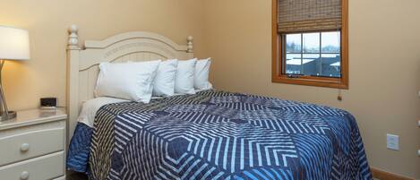 A bedroom with a bed, a nightstand, a lamp, a window with blinds, and a view of buildings outside.