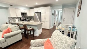 A modern kitchen with white cabinets, stainless steel appliances, and a central island. There is a living area with patterned sofas and a dining table. - Carolina Beach Ocean View Retreat w/ Beach Access! (Carolina Beach)