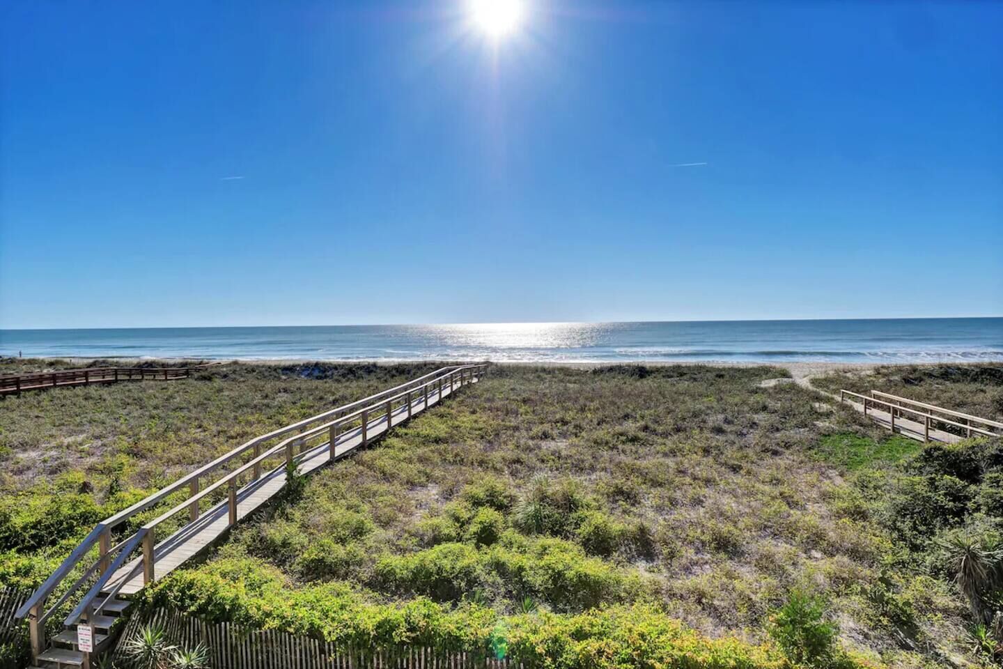 Una pasarela de madera que lleva a una playa, con vista despejada al océano y un cielo soleado.
