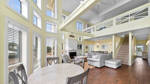 A spacious living area with a high ceiling, a fireplace, a dining table, and a staircase leading to an upper level. - Calypso Breeze | Gulf Views (Orange Beach)