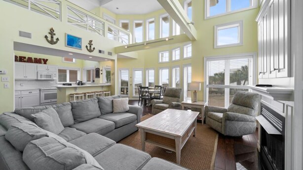 A spacious living area with a large gray sectional sofa, a wooden coffee table, and a fireplace. There is a kitchen area with white cabinets and a dining table with chairs. The room has large windows and a balcony with railings. - Calypso Breeze | Gulf Views (Orange Beach)