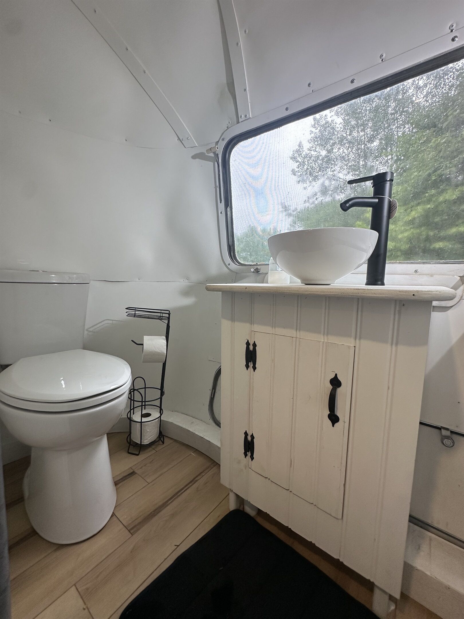 Een kleine badkamer met een wit toilet, een witte wastafel met een zwarte kraan, en een raam boven de wastafel.