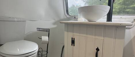 Een kleine badkamer met een wit toilet, een witte wastafel met een zwarte kraan, en een raam boven de wastafel.