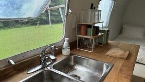 Eine kleine Küche mit Spüle aus Edelstahl, einem Fenster mit Blick auf eine Rasenfläche und einem Regal mit Küchenutensilien.