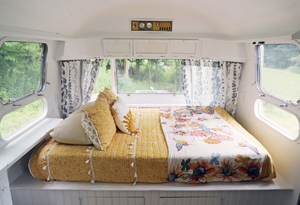 A cozy bed with patterned bedding and pillows, a window with curtains, and a small shelf above the bed.