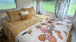 Ein Bett mit floralem Überwurf und zwei Kissen, ein Fenster mit Vorhängen und ein Holzboden.