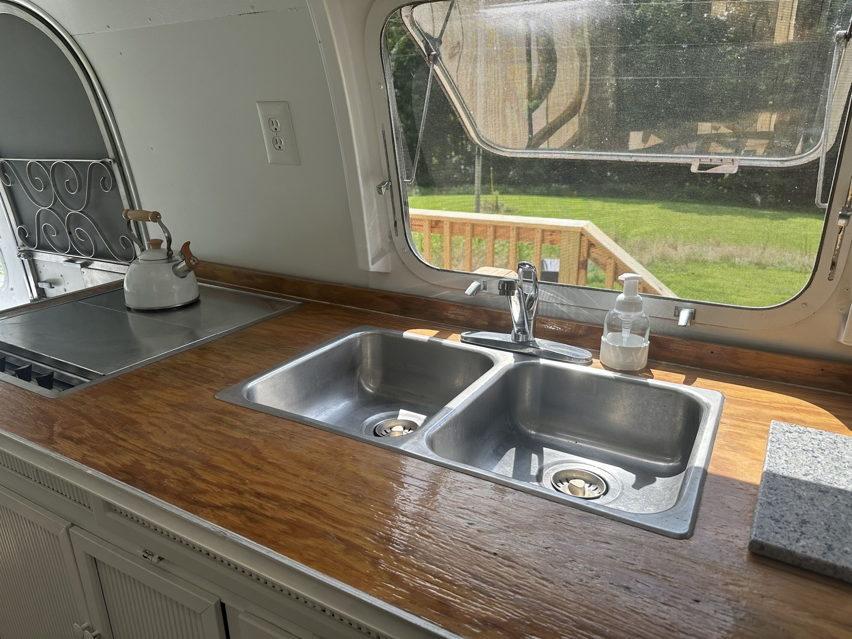 Eine Küche mit doppelter Spüle, Wasserkocher und Fenster mit Blick auf eine Holzterrasse.