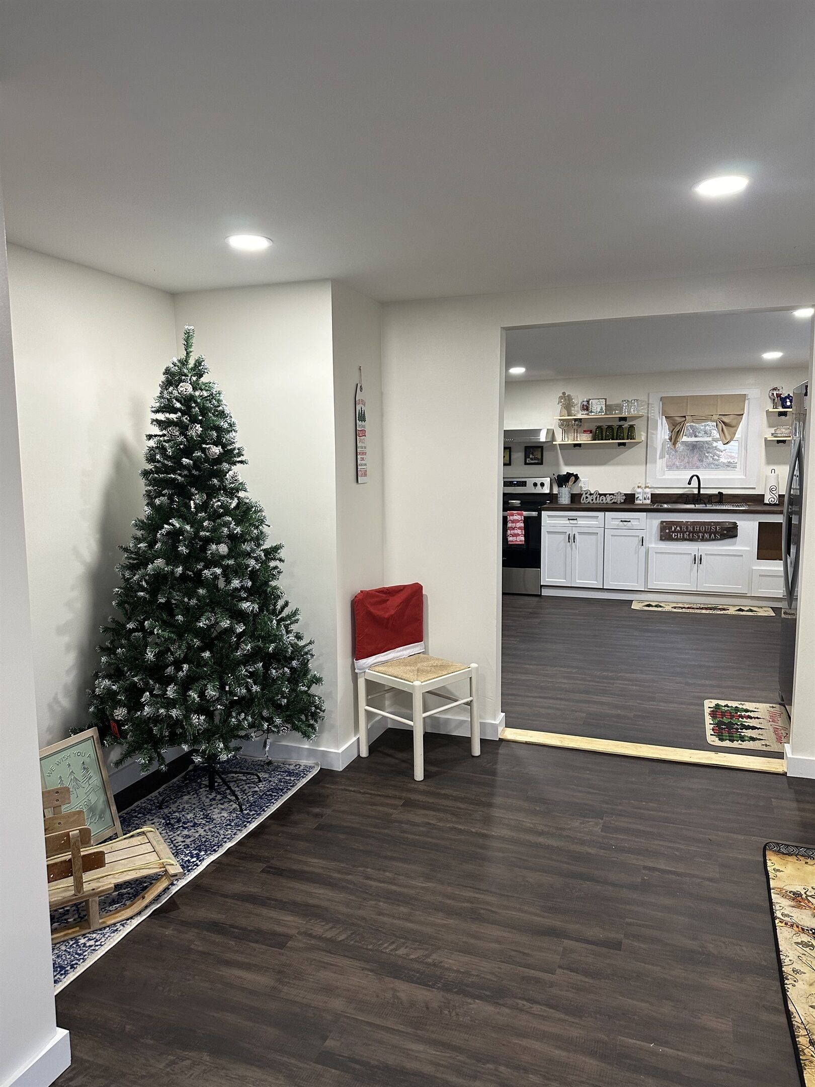 Un salon moderne avec un parquet en bois foncé, un fauteuil blanc, un petit sapin de Noël et une cuisine visible à travers une porte ouverte.