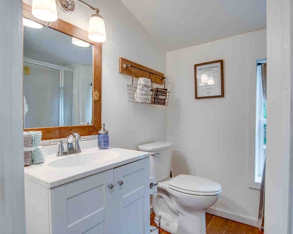 A bathroom with a white sink, mirror, and toilet. There is a towel rack above the toilet and a framed sign on the wall.
